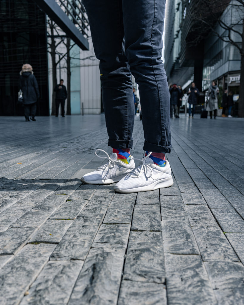 Brightly coloured socks stand out in the middle of the frame. Beneath them bright white trainers stand on grey brick pavement, Above them, legs in dark jeans ascend out of the frame.