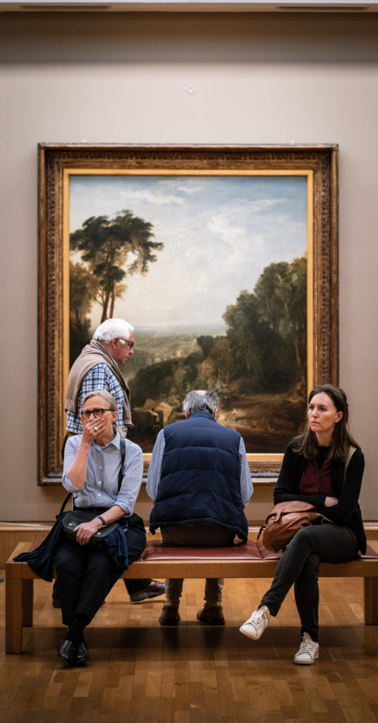 Gallery attendees sit and walk bay a Turner's 'Crossing the Brook', which dominates the wall behind them. All the people look bored. One stares into the middle distance while another covers a yawn with her hand.