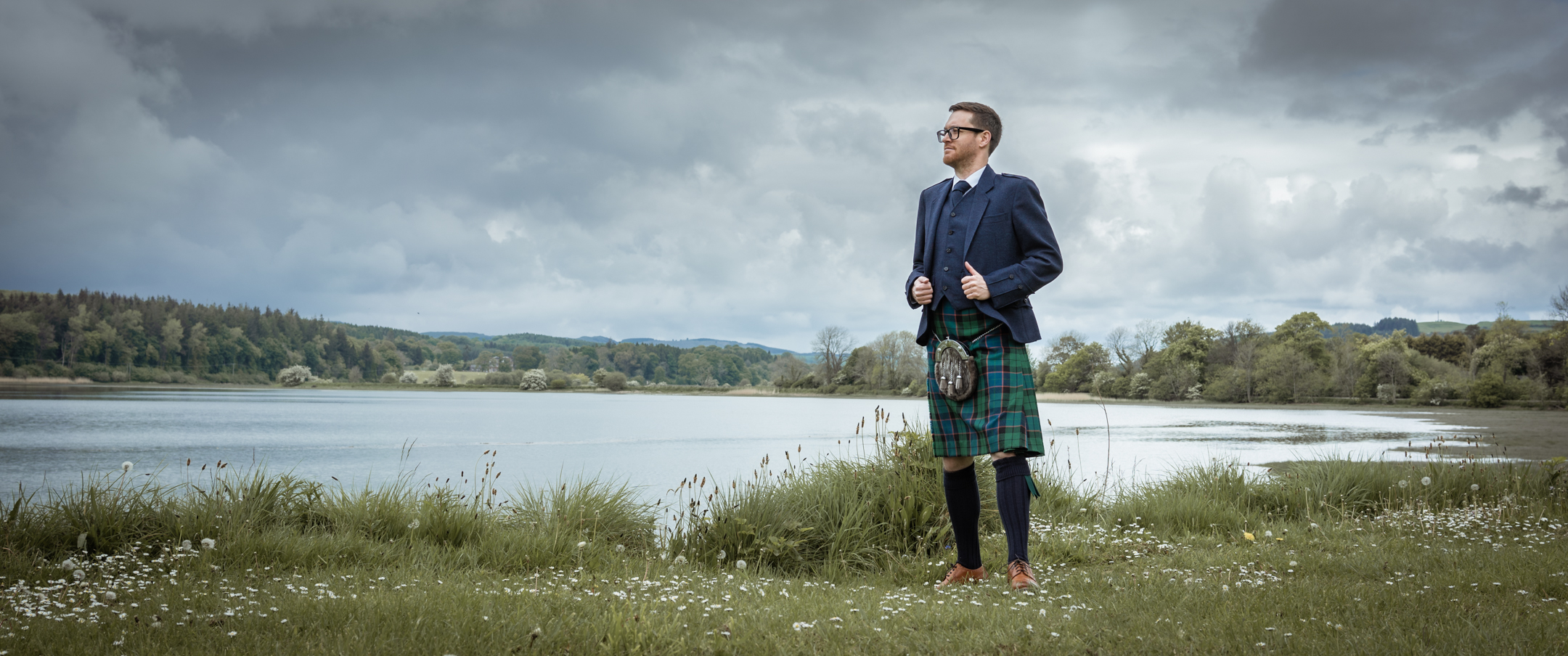 A man stands in a green and blue kilt with a blue jacket and waistcoat against a backdrop of a wide river, cloud-filled sky and wild greenery.