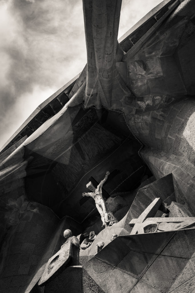 An angular sculpture of Christ, lit by strong afternoon sun, hangs starkly against the darker, muscular architecture of the Sagrada Família's Passion Façade.