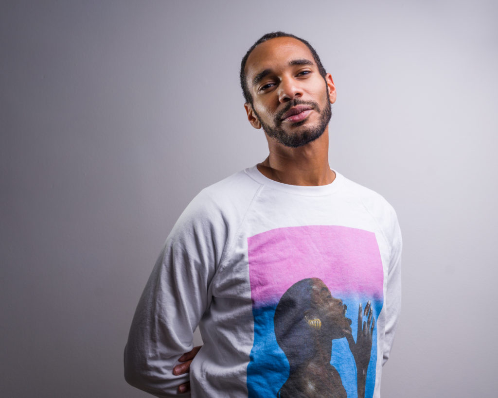 Studio portrait: Adam wears a white sweatshirt with a bold print of a dark skinned woman against a vivid pink and blue background. He is standing proud but relaxed, with his arms folded casually behind his back.