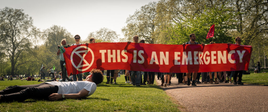 A man lying on the grass has raised his head to look behind him, where a protest march bears down on his location. The protesters are holding a large red banner emblazoned with the slogan 'This is an emergency' and the Extinction Rebellion logo.