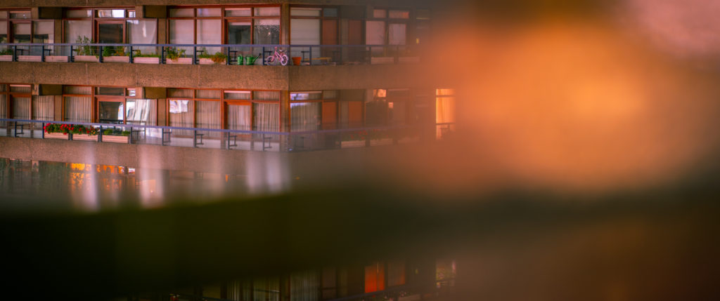 The balconies and honeyed wooden window frames of a concrete residential building blend with abstract shapes created from out-of-focus objects oranges and dark green.