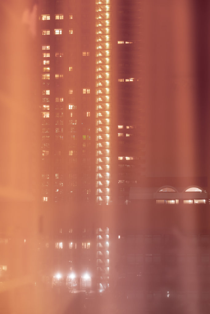 The lights of a residential tower block at night make a grid-like pattern in a diffuse, indistinct scene, completely shot through with reddish hues and low contrast.