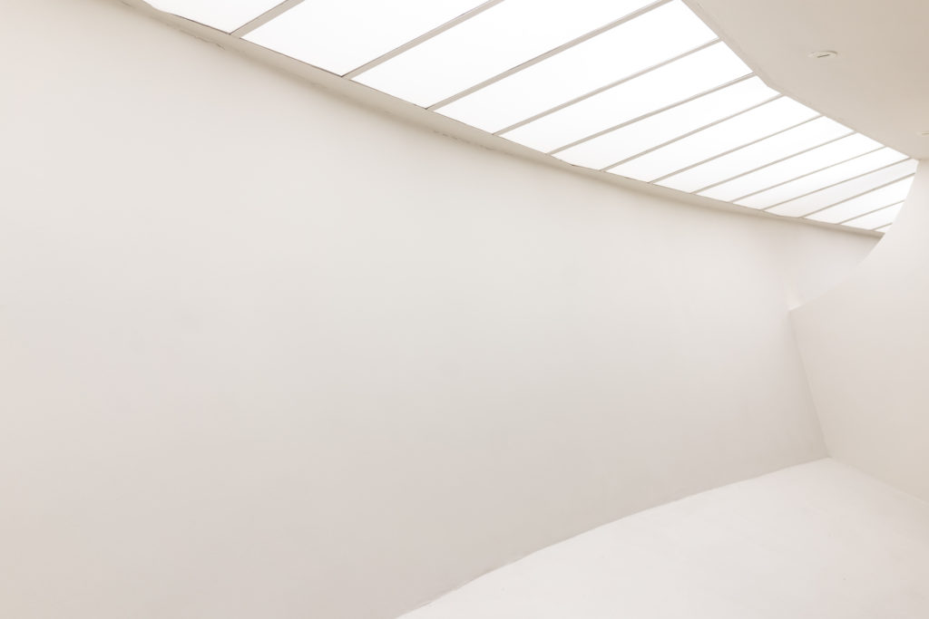 Architectural details in a completely white room with curved walls meeting at various angles and a row of light panels in the ceiling.