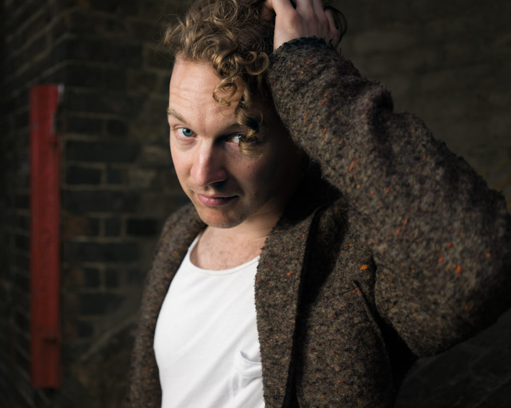 A man stands for his portrait in front of a dark, brown-brick wall. He wears a white t-shirt and brown jacket, which is shot through with flecks of terracotta and orange. His hand is in his hair, which partially hangs in front of his face.