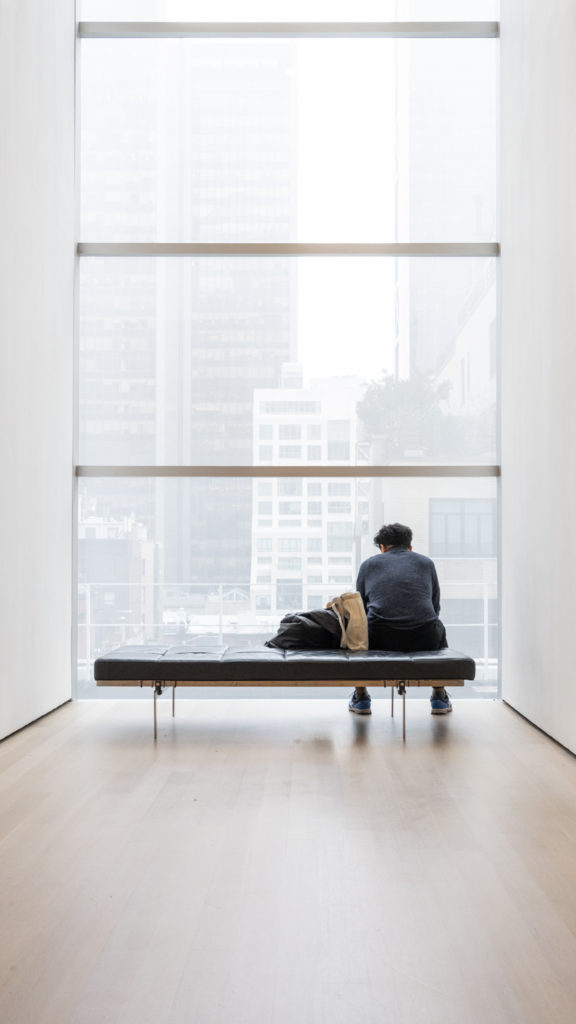 A man sits on a bench tucked in a quiet corner of a gallery, before him a giant window revealing hints of the New York cityscape beyond. The room is white, with pale wood flooring, punctuated by the stark contrast of the person, their belongings and the bench, all of which are dark.