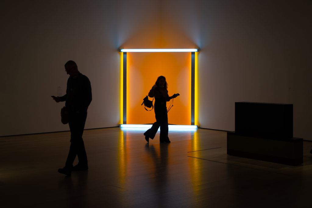 A light-based artwork stands in the corner of a gallery room, taking the form of a square frame with white lights at the top and bottom, yellow lights at the sides and an orange glow in the space behind. A woman walks in front of it, silhouetting her mid-stride. Another man is walking away, almost enveloped in darkness, glancing at his phone.