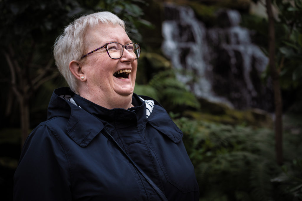 My mum laughing in front of dark tropical plants and a waterfall.