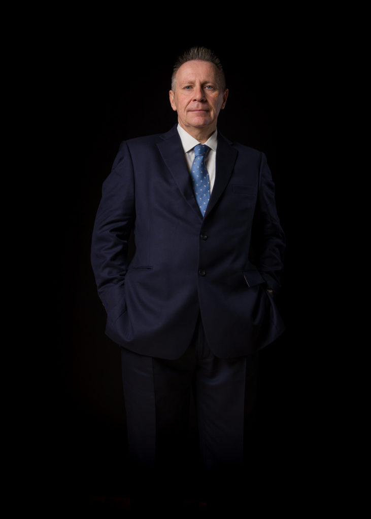 Lord Guy Black of Brentwood, in a deep navy suit against a dark background, stands purposefully but approachably, hands in pockets