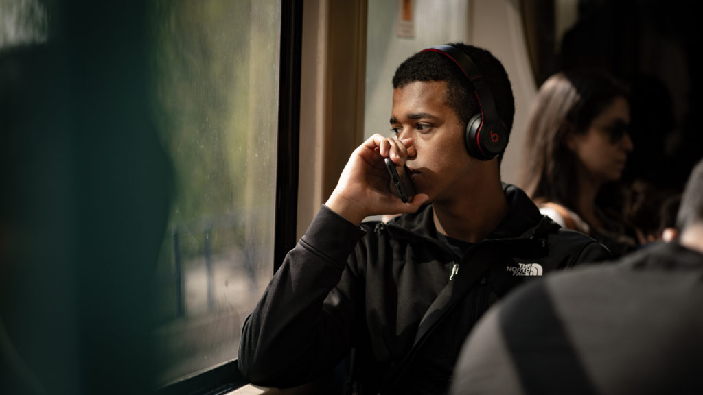 A man on a train looks out of a large window, his elbow resting on the frame, supporting the hand he holds up to his face. His phone is in his hand, its edge pressed against his lips. He is lost in thought as his headphones pipe music into his ears. The scene is warm and summery.