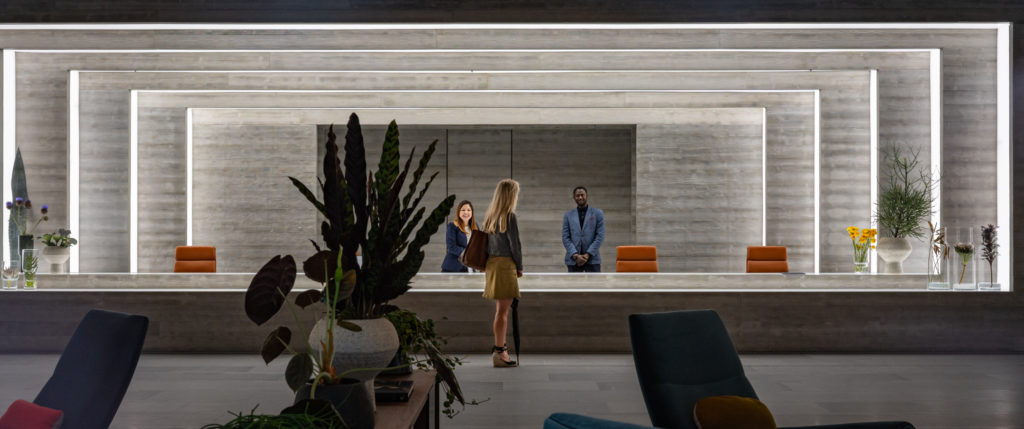 A woman stands at a very wide reception desk and two members of staff stand on the other side, smiling at her. The reception area is made of increasingly recessed rectangular concrete arches, each one rimmed with light panels. The room is dotted with designer furniture and plants.