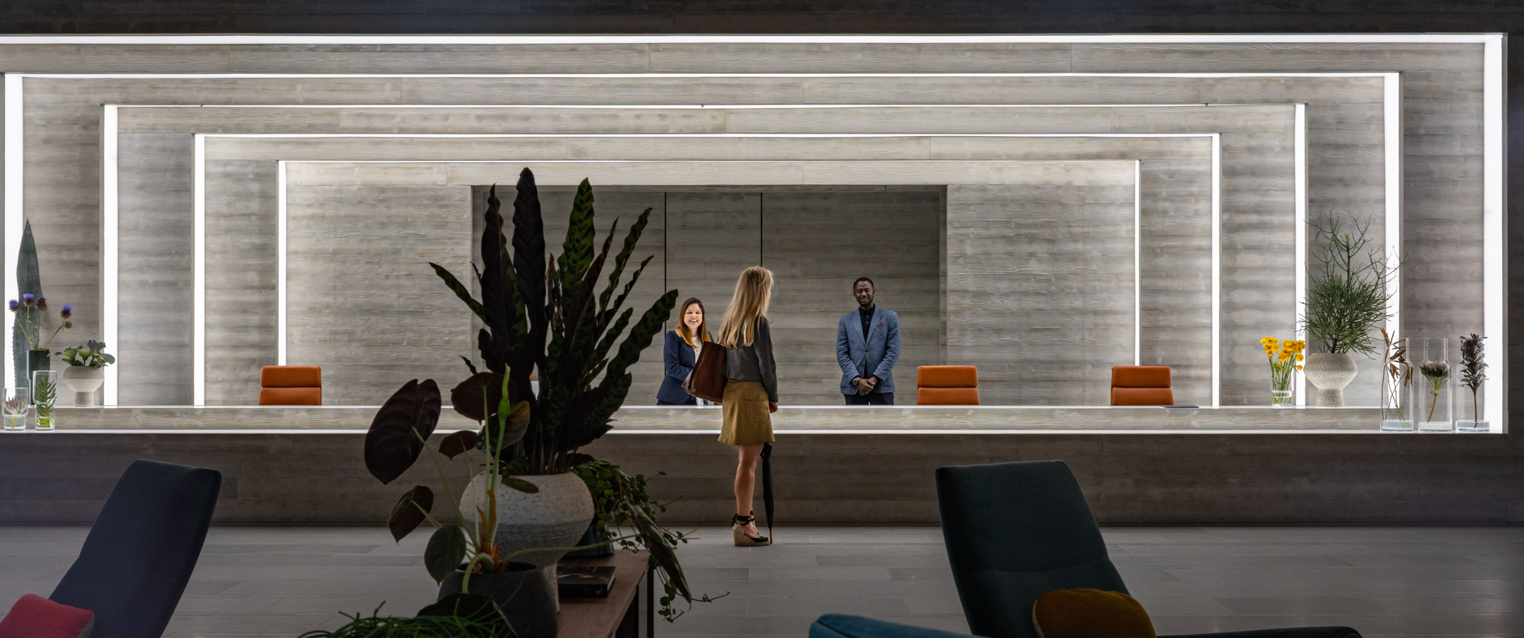 A woman stands at a very wide reception desk and two members of staff stand on the other side, smiling at her. The reception area is made of increasingly recessed rectangular concrete arches, each one rimmed with light panels. The room is dotted with designer furniture and plants.
