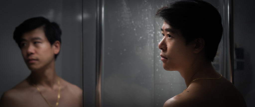 A man with his back to the camera stands in front of a bathroom mirror. His face is softly lit and most of the rest of the room falls into semi-darkness. Water droplets on a shower screen glint behind his head. He is topless wearing a fine gold chain. He looks to the side, revealing the light side of his face in his reflection.