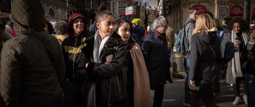 Two women, one with folded arms, stand fast amid a crowd moving in all directions, both watching something out of frame.