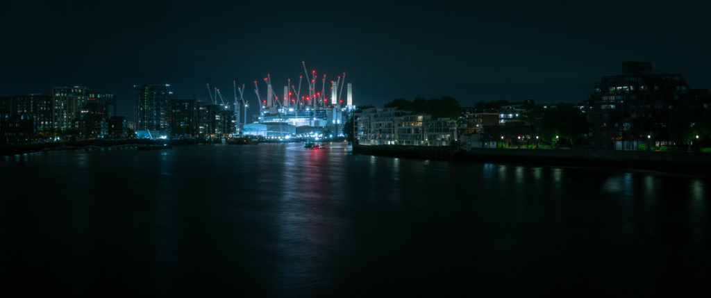 Looking along the River Thames on an inky-black night towards Battersea Power Station, which glows bright with intense cool tones crowned by a swarm of red warning lights. The comparatively weak light of buildings shouldering the river provide only dim suggestions of their shapes.