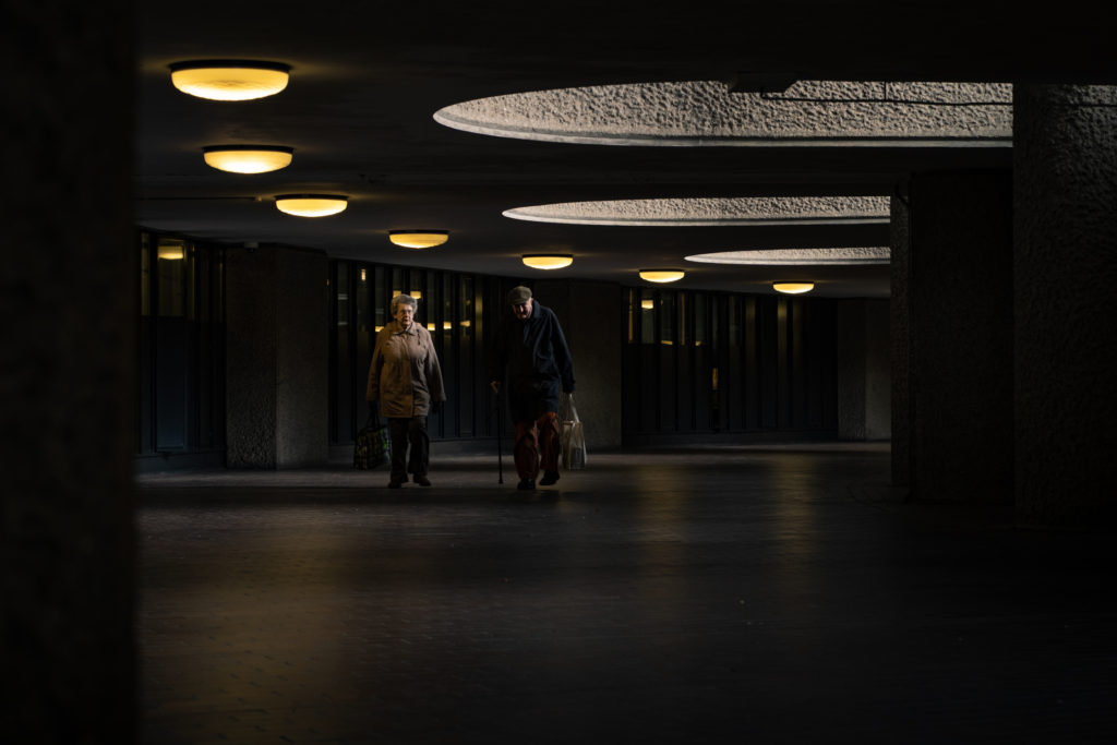 A dark brutalist crescent lit by an arc of yellowish lights and sunlight coming through large ceiling recesses plays host to two people on their way home from a shopping trip.