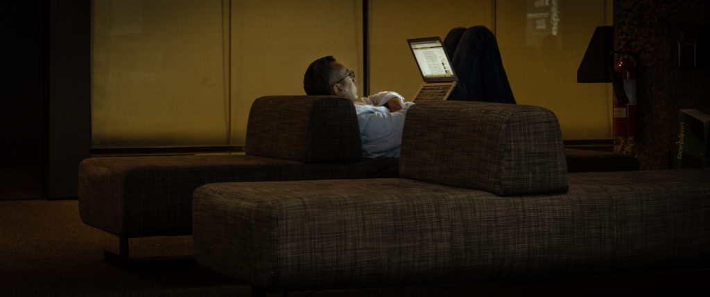 A man in a light blue shirt reclines on a low seat, his arms folded and knees hitched up supporting an open laptop. The screen lights his face and front radiantly in clean light while glass panels behind glow murkily in a deep yellow-green hues. The rest of the scene falls gently into darkness.