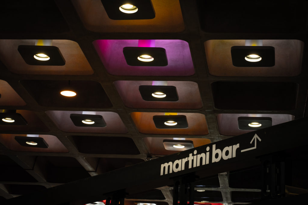 A thick black guardrail cuts boldly in a diagonal line across the lower third of the picture. It carries bold white lettering in Futura and a sharply angled arrow, which indicate a Martini Bar upstairs. Above, the polished concrete ceiling is pocked with checkerboard recesses, some empty, some containing neutral downlighters - themselves backlit in saturated oranges and purples.