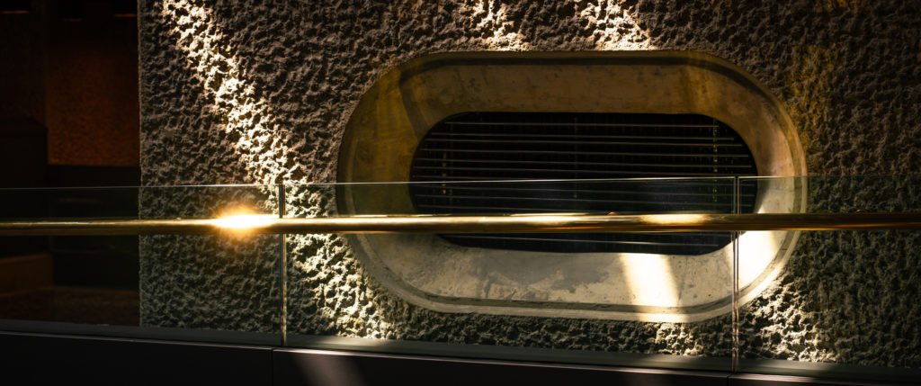 An air vent - expertly crafted as a perfect geometric stadium - sits recessed into a wall, which carries a hammered concrete finish. A brass handrail bisects at an odd angle as it follows the line of a gently descending walkway. Sunlight breaks through a skylight, streaks across the wall in shafts and causes the handrail to glisten and flare.