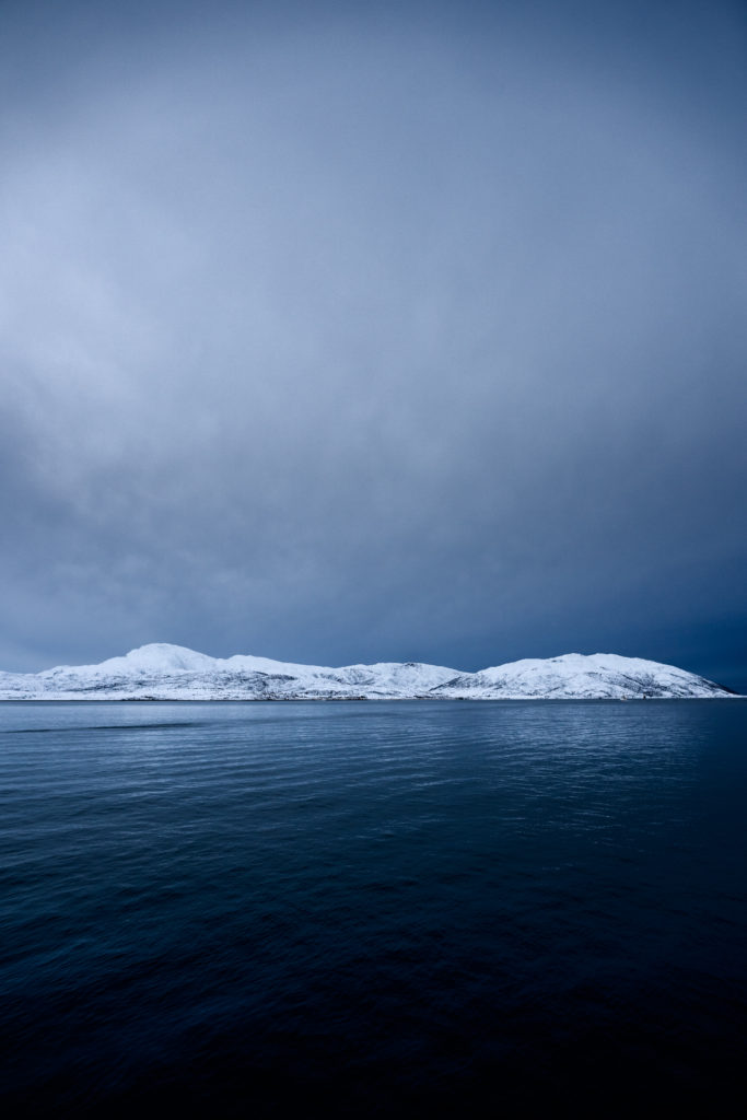 Snowy hills sandwiched between still, deep blue fjord water and an atomspheric sky.