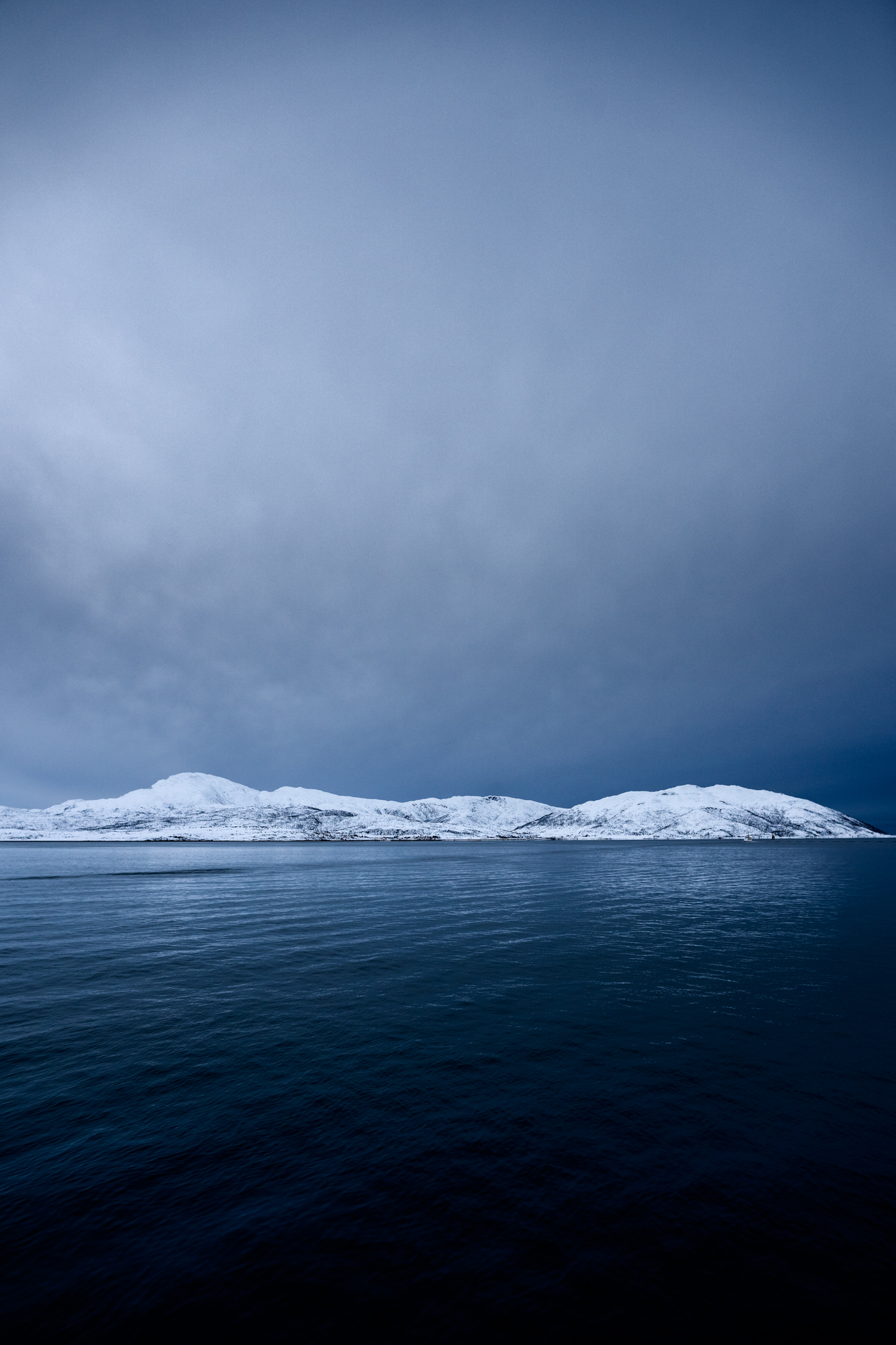 Snowy hills sandwiched between still, deep blue fjord water and an atomspheric sky.