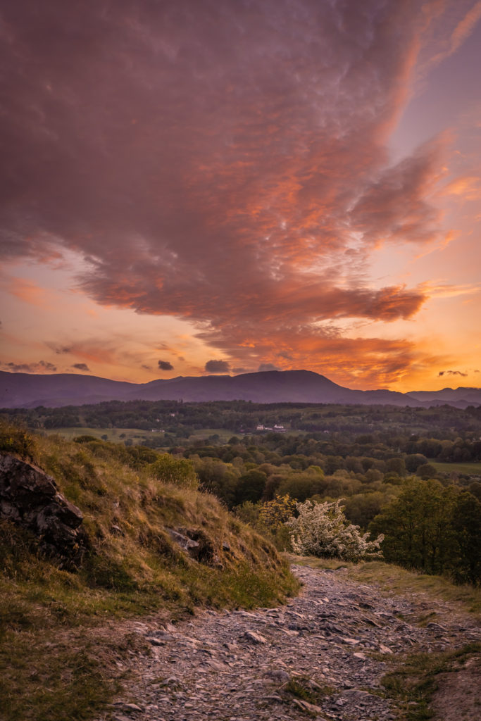 A rough stone path winds behind a grassy slope in the foreground, mirroring a long train of firey orange clouds stretching over a tree-dotted valley, beyond the mountains on the horizon and into the heart of the sunset sky.