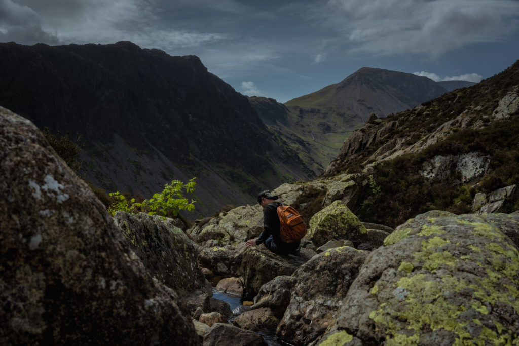 A lone figure, in a black baseball cap with a burnt orange rucksack, sits on large boulders on the slopes of Fleetwith Pike, water trickling past, as the craggy greys, browns and greens fold over each other into a spectacular mountain scene.