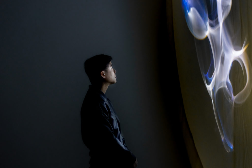 Low key, side portrait of a man who is lit entirely by abstract lighting patterns in Olafur Eliasson's Eine Beschreibung einer Reflexion, oder aber eine angenehme Übung zu deren Eigenschaften, which towers on the right-hand side of the frame.